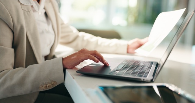 A business woman's hands typing on a laptop while holding a printed document