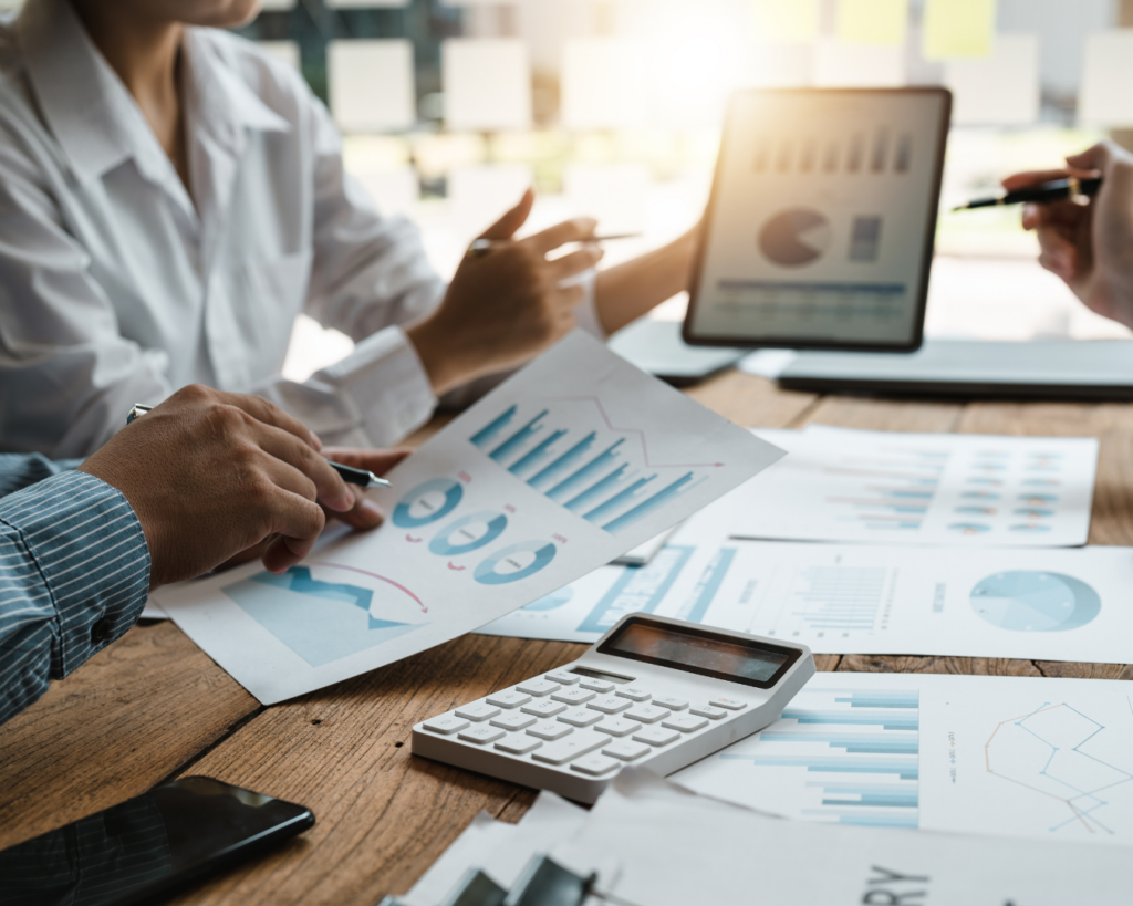 A bright and airy photo of documents filled with charts and graphs strewn about a table top. Several people's hands are visible gesturing towards figures, and one person holds a tablet displaying more charts and graphs.