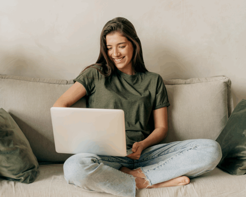 A young woman sits cross-legged on a couch with a laptop on her knee, smiling.